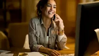 Mulher sorri enquanto fala ao celular sentada em frente à tela do computador