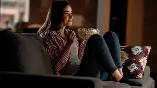 Mulher sorri sentada no sofá com um balde de pipoca na mão enquanto se diverte vendo TV